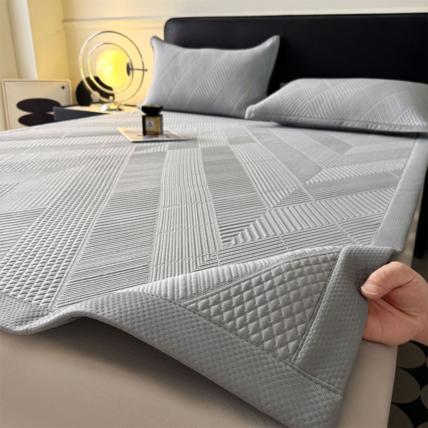 A light gray quilted bed sheet with a geometric patterned design is shown on a bed, with a hand lifting the corner to demonstrate its thickness and texture. The bed is styled with matching gray pillows and a dark headboard.