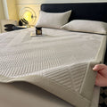 A quilted bed sheet with a geometric diamond pattern in light beige and off-white, shown on a bed with matching pillows. A hand is visible in the bottom right corner holding the corner of the sheet to demonstrate its drape and texture.