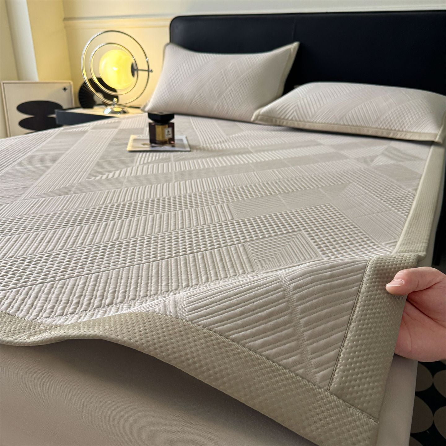 A quilted bed sheet with a geometric diamond pattern in light beige and off-white, shown on a bed with matching pillows. A hand is visible in the bottom right corner holding the corner of the sheet to demonstrate its drape and texture.
