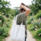 A woman standing on a garden path carrying a large white canvas tote bag with a circular white logo patch on the front, photographed outdoors in a blurred green garden setting.