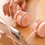 A close-up of a person's hand adjusting a small pink portable speaker connected to a smartphone on a wooden table. Another matching pink speaker is visible nearby.