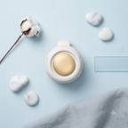 A round white cosmetic compact with a gold-toned circular button, styled in a flat lay on a light blue textured background with cotton stems, marble stones, and a folded gray cloth.